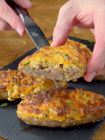 A hand uses a knife to lift a slice of cooked ground meat patties topped with melted cheese and vegetables from a dark serving board. Other cheesy patties are visible in the background.