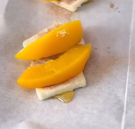 Two slices of peach placed on top of a piece of white cheese with honey drizzled underneath, all sitting on a sheet of parchment paper.