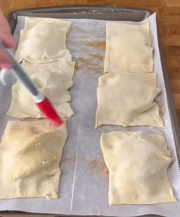 A hand brushes egg wash onto unbaked pastry pockets arranged on a parchment-lined baking sheet, preparing them for baking.