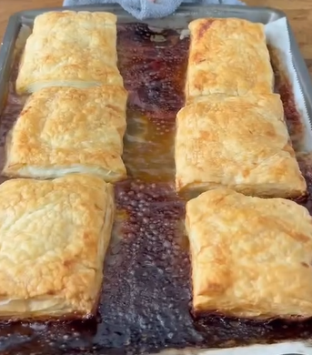 Six golden-brown puff pastry squares are arranged in two rows on a baking sheet, with some caramelized filling oozed and baked onto the pan between them.