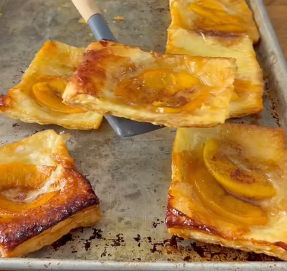 Rectangular peach pastries with golden, flaky crusts are arranged on a metal baking sheet. One pastry is being lifted with a spatula, showing its caramelized peach slices and glossy surface.