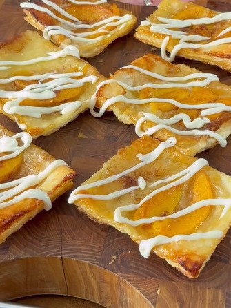 Six square peach pastries with golden puff pastry, topped with peach slices and drizzled with white icing, displayed on a wooden board.