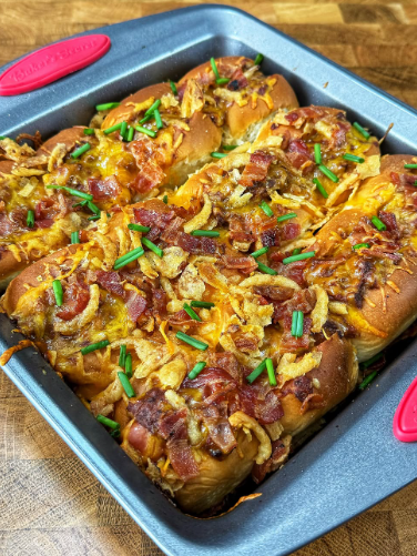 A square baking pan filled with baked rolls topped with melted cheese, crispy bacon pieces, fried onions, and chopped chives, sitting on a wooden surface.