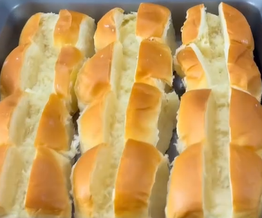 Four rows of freshly baked, golden brown bread rolls are placed closely together in a baking tray, each row featuring soft, fluffy interiors visible from the side.