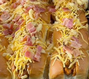 Hot dogs topped with shredded cheddar cheese, crispy bacon bits, and crunchy fried onions, served in buns and arranged side by side.