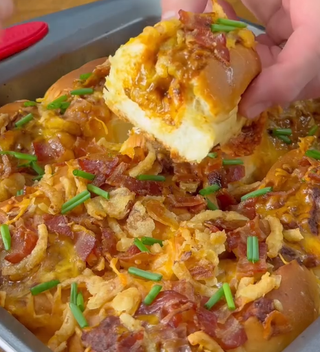 A hand lifts a cheesy, bacon-topped slider from a tray of baked pull-apart rolls, garnished with chives and crispy onions.