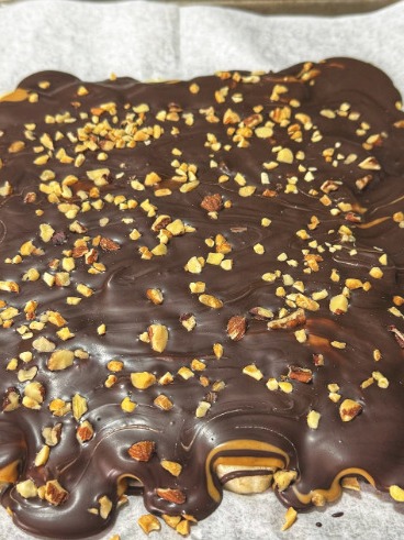 A square dessert covered in glossy melted chocolate and sprinkled with chopped nuts, resting on a sheet of parchment paper. Some caramel is visible under the chocolate along the edges.