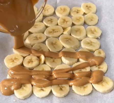 Sliced bananas arranged on parchment paper with creamy peanut butter being poured over them from a bowl.