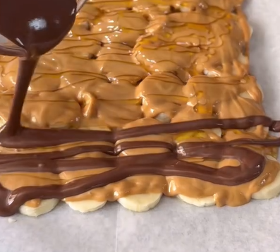 Chocolate is being drizzled over a layer of peanut butter and sliced bananas arranged on a rectangular base, all placed on parchment paper.