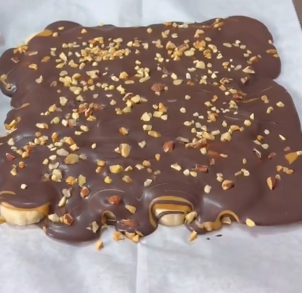 A slab of chocolate-covered caramel with chopped nuts sprinkled on top, set on a sheet of parchment paper.