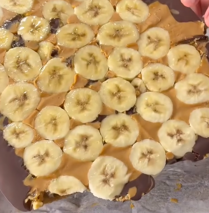 A close-up of a dessert topped with banana slices, creamy peanut butter, and a chocolate layer on a baking sheet. A hand is visible in the corner, holding the edge of the dessert.