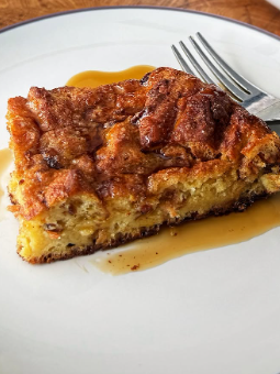 A slice of bread pudding sits on a white plate with a fork beside it, drizzled with syrup.