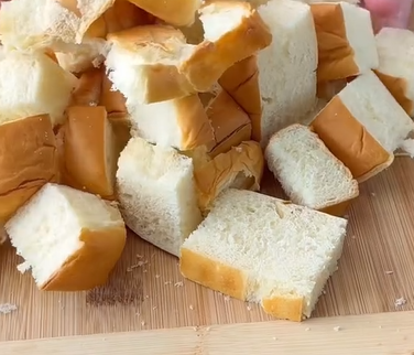 Cubed pieces of white bread are piled on a wooden cutting board, ready for use in a recipe or dish.