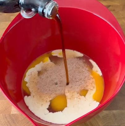 A close-up of a red mixing bowl with melted butter and sugar inside, while a dark liquid, possibly vanilla extract or cola, is being poured in from a bottle above. The bowl sits on a wooden surface.