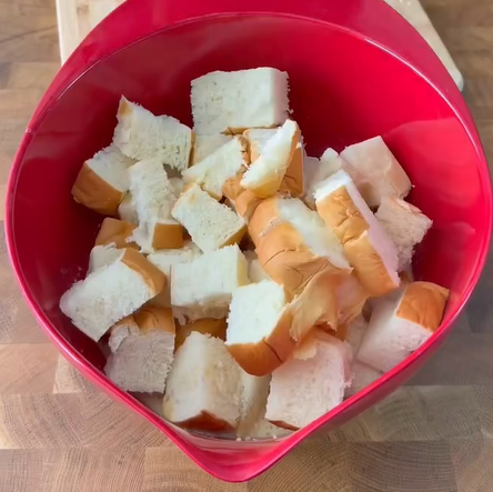 A red mixing bowl filled with cubed pieces of white bread sits on a wooden countertop.