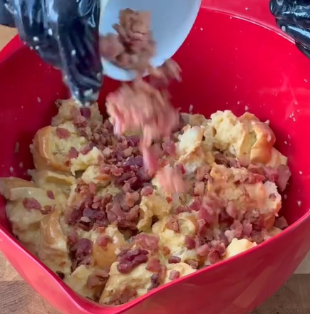 A person wearing black gloves pours chopped bacon into a large red bowl filled with bread pieces and a creamy mixture, preparing a savory dish on a wooden surface.