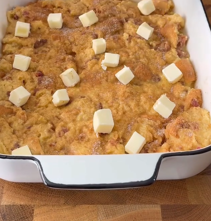 A white rectangular baking dish filled with unbaked bread pudding, topped with evenly spaced cubes of butter, sits on a wooden surface.