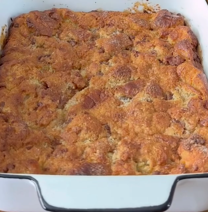 A close-up of a golden-brown bread pudding in a white rectangular baking dish, showing a textured, slightly crispy top with chunks of bread visible.