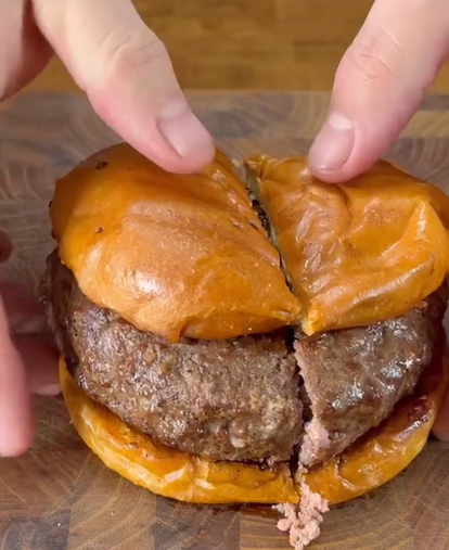 A close-up of hands pulling apart a burger with a thick beef patty on a toasted bun, sitting on a wooden surface. The burger is sliced in half, revealing the juicy patty inside.