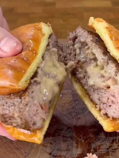 A close-up of hands holding a burger sliced in half, revealing melted cheese oozing from the center of the beef patty inside a soft, golden bun.