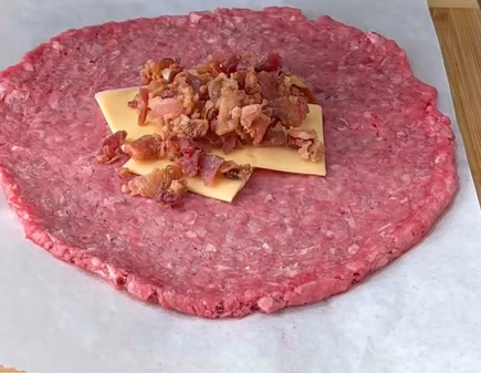 A large, flat circle of raw ground beef on parchment paper with two slices of cheese and a heap of cooked, crumbled bacon placed in the center.