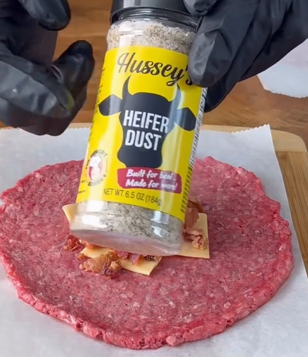 A gloved hand sprinkles Husseys Heifer Dust seasoning onto a layer of raw ground beef topped with cheese and bacon pieces, preparing a meat dish on parchment paper.