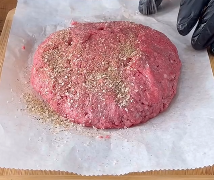 A mound of raw ground beef seasoned with salt and pepper sits on parchment paper, with a gloved hand visible on the right side, preparing the meat mixture.