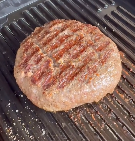 A thick beef burger patty with grill marks is cooking on a black grill grate.