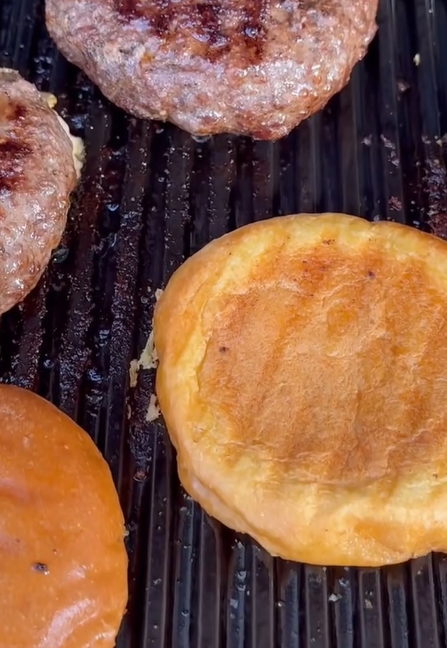 Burger patties and golden brown burger buns are being grilled on a ribbed grill surface, showing grill marks and a slightly oily texture.