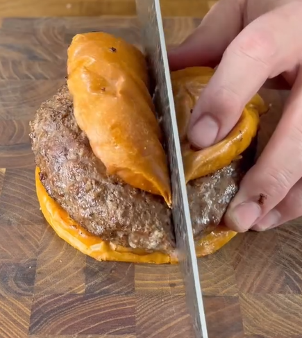 A hand slices a hamburger in half on a wooden cutting board. The burger features a thick beef patty and a shiny, golden-brown bun.