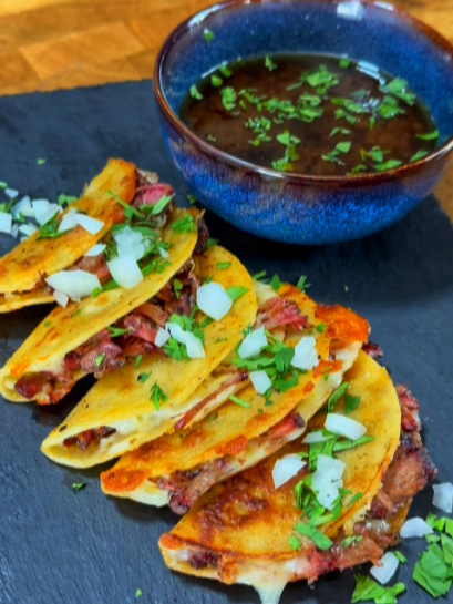 Three birria tacos filled with meat, topped with chopped onions and cilantro, are arranged on a slate plate next to a blue bowl of consommé garnished with fresh herbs.