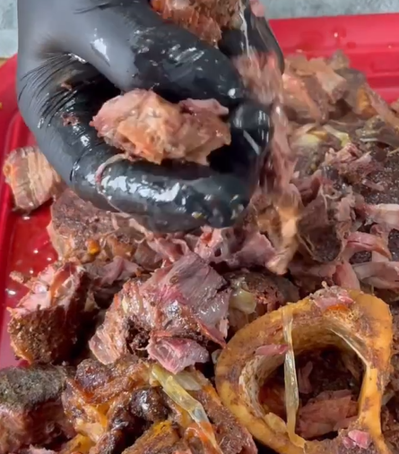 A gloved hand holds a chunk of shredded, cooked meat over a pile of seasoned meat and a large bone on a red cutting board.