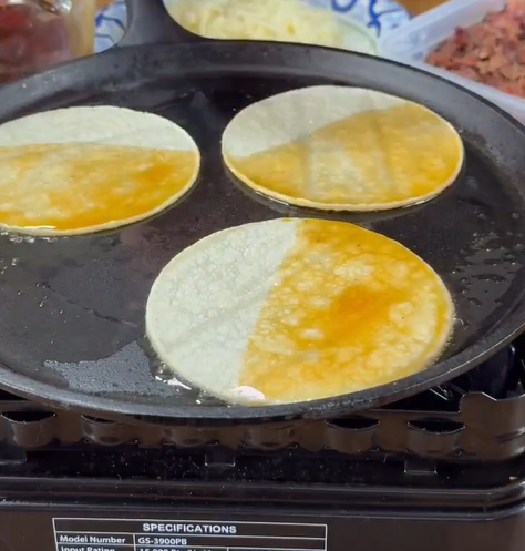 Three corn tortillas are heating on a flat griddle, with one half of each tortilla covered in a layer of orange sauce or oil. Ingredients are visible in containers in the background.