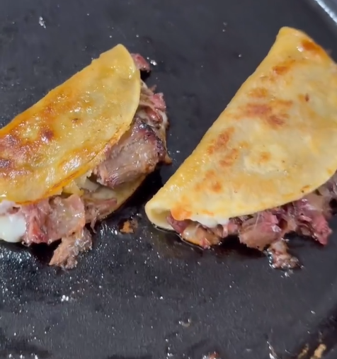 Two tacos with crispy, golden-brown corn tortillas are filled with shredded meat and melting cheese, cooking on a hot, slightly greasy skillet.