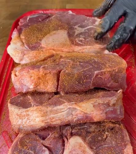 Four raw steaks on a red cutting board are being seasoned with dry rub by a person wearing a black glove.