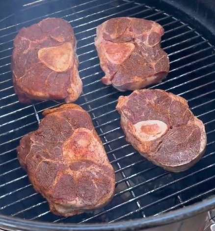 Four thick, seasoned cuts of meat with bones in the center are grilling over a barbecue, cooking on a metal grate. Smoke is visible, suggesting the meat is being slow-cooked or smoked.