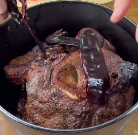 A close-up of a cooked, seasoned beef shank in a black pot, with two rehydrated dried chili peppers draped over the meat and hands adjusting the chilies.