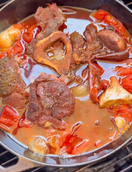 A pot of stew with chunks of meat, beef bone marrow, red tomatoes, and onions simmering in a rich, reddish broth.