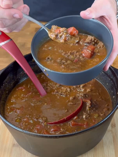 A person holds a bowl of chunky beef stew with tomatoes above a pot filled with more stew, using a ladle and a spoon to serve the dish.