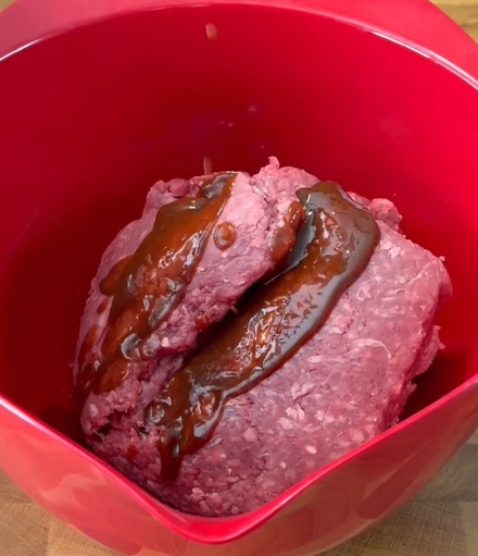 Ground beef in a red mixing bowl with a drizzle of brown sauce on top, ready for mixing.