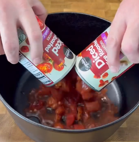 Two hands pour canned diced tomatoes into a black pot on a wooden surface. The cans are labeled Diced Fire Roasted Tomatoes.