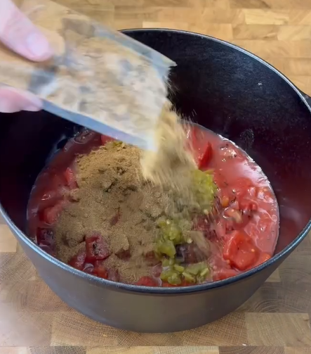 A hand pours a packet of brown seasoning into a black pot filled with diced tomatoes and chopped green chilies, on a wooden surface.