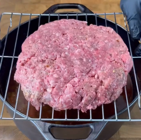 A large, uncooked ground beef patty sits on a wire rack over a black cast iron pot. A person wearing a black glove is visible on the right edge, preparing the meat for cooking.