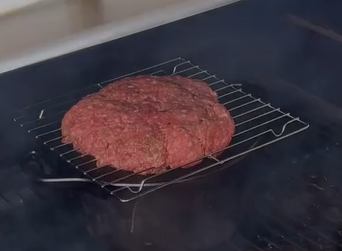 A large, uncooked hamburger patty sits on a wire grill rack over a stove or grill, with smoke rising around it.