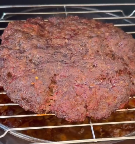 A large, well-cooked piece of ground meat sits on a metal grill rack, with juices dripping into a pan below. The meat has a dark, crispy exterior.