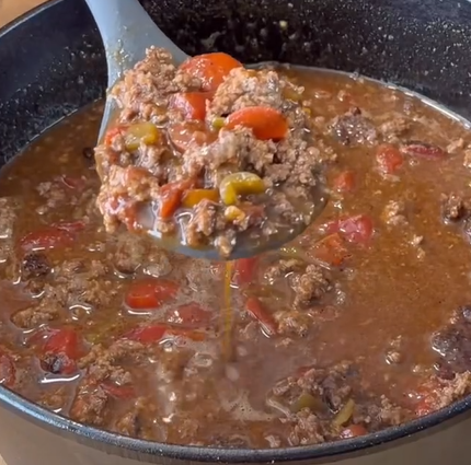 A close-up of a ladle lifting a serving of chili with ground beef, diced tomatoes, and green peppers from a black pot filled with the hearty, chunky stew.