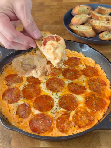 A hand dips a toasted bread slice into a skillet of hot, cheesy pepperoni dip. A plate of toasted bread slices sits in the background on a wooden surface.