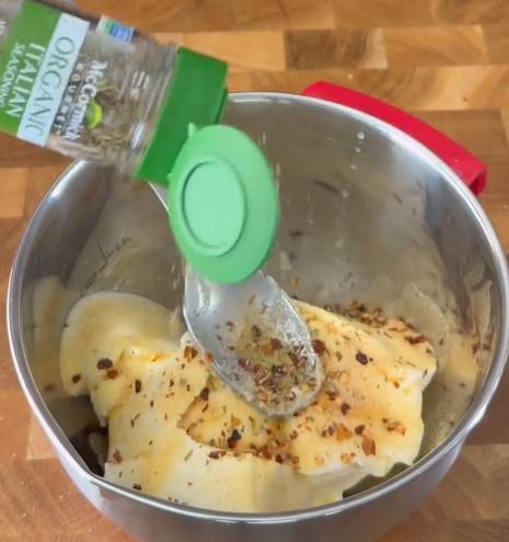 A metal mixing bowl contains lasagna noodles with cheese and spices. A hand sprinkles Italian seasoning from a green-capped jar over the mixture. A spoon rests inside the bowl on a wooden surface.