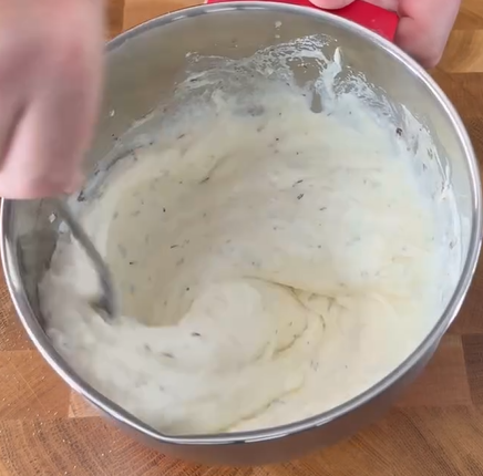 A hand stirs a thick, white mixture with herbs in a stainless steel bowl on a wooden surface.