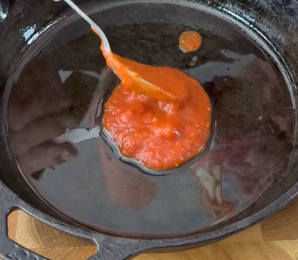 A spoon is spreading tomato sauce in the center of a black cast iron skillet on a wooden surface.